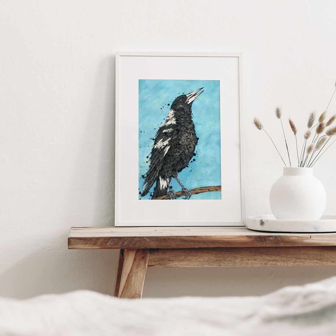 Shannon Dwyer fine art print featuring an artwork of ‘Carol’ the Australian Magpie bird with black and white feathers, perched on a branch, with a splash of blue in the background. Framed on a sideboard.